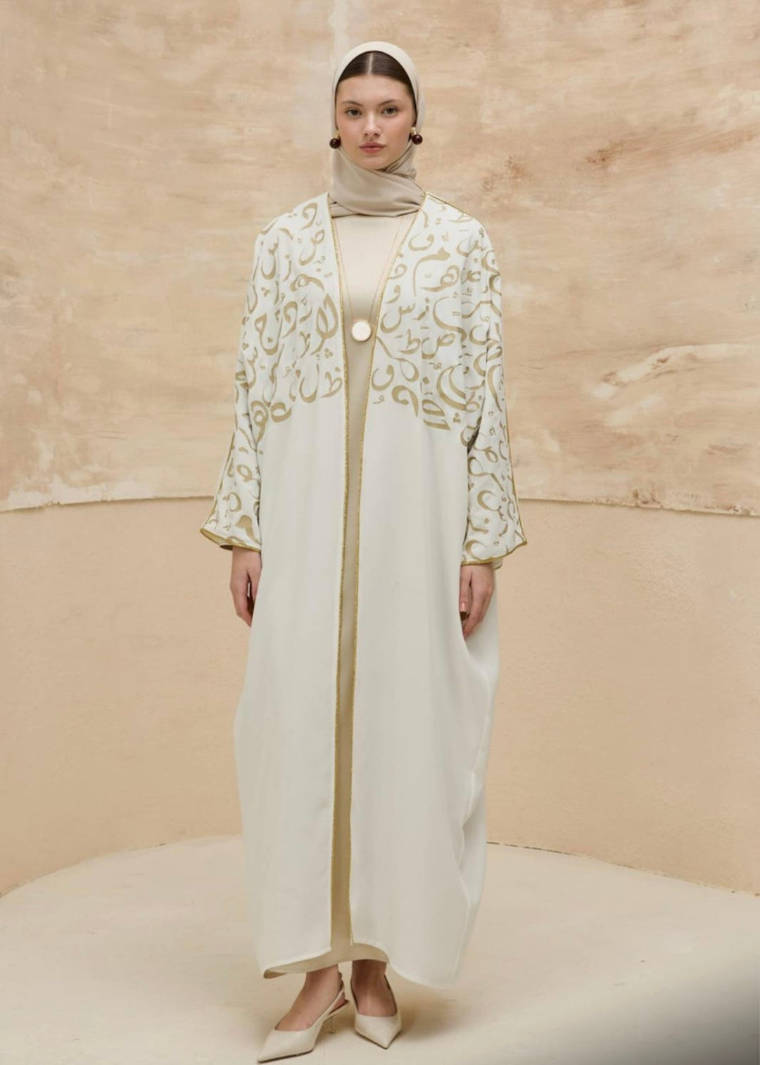 Woman wearing a long, embroidered white dress with a matching headscarf against a beige background.